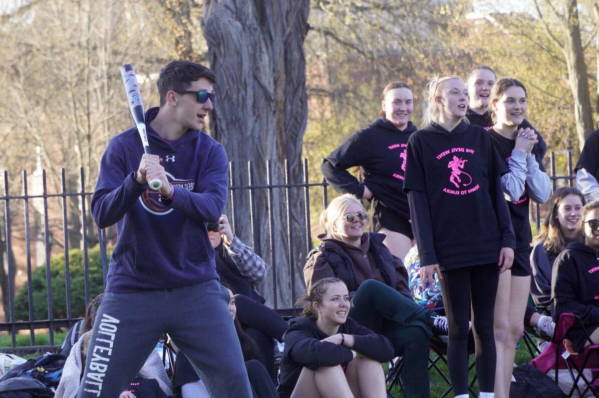 A male student gets ready to hit during Term III softball as a group of female students look on behind him